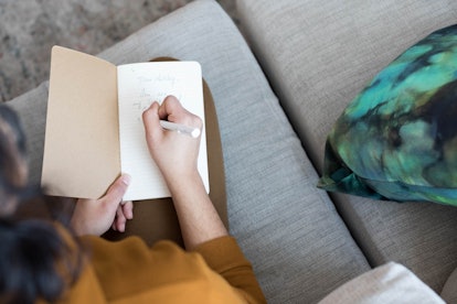 A brunette woman sitting on a grey couch writing something down in a journal