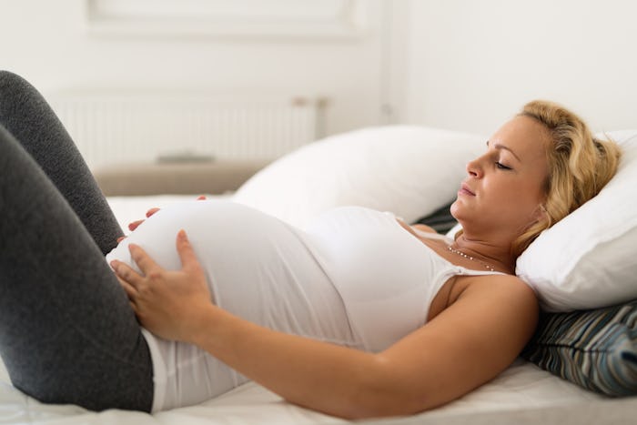 pregnant woman laying on back in white bed looking down holding belly uncomfortably