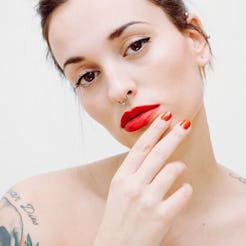 A woman with red false nails, a tattoo on her shoulder, her hair in an updo wearing a red lipstick 