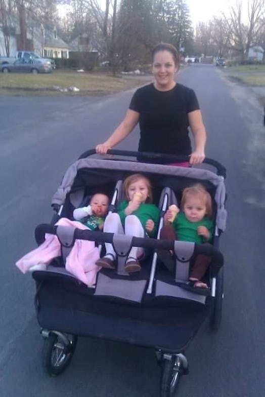 A woman is pushing her three kids in a baby stroller.
