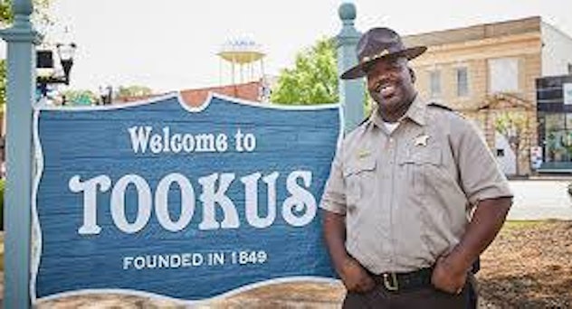 A man posing next to a "Welcome to Tookus founded in 1549" blue sign