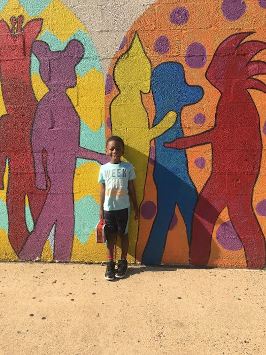 A smiling child posing next to a painted wall, whose mom has back-to-school anxiety that white moms ...