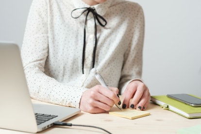A woman writing in a sticky note with a laptop in front of her and her notebook and phone beside her