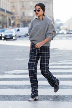 A brunette woman crossing a street in a grey cozy chunky knit fall sweater, check pants, and grey bo...