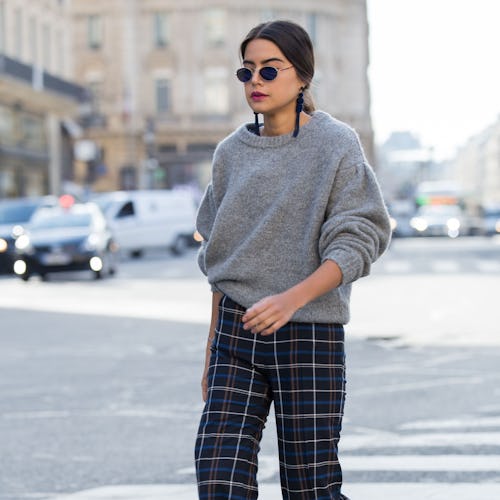 A brunette woman crossing a street in a grey cozy chunky knit fall sweater, check pants, and grey bo...