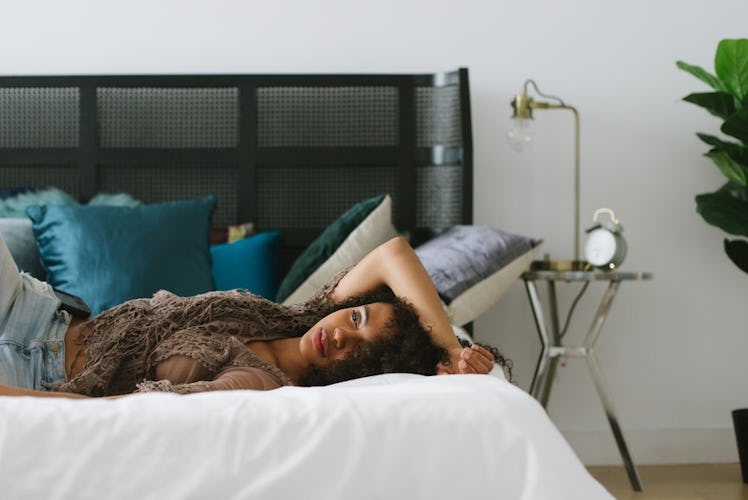 A woman in a brown shirt, and blue denim pants lying on her bed with her AC on