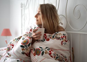 A woman on her period, lying in her bed covered with a floral duvet while holding a white mug, feeli...