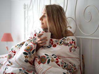 A woman on her period, lying in her bed covered with a floral duvet while holding a white mug, feeli...