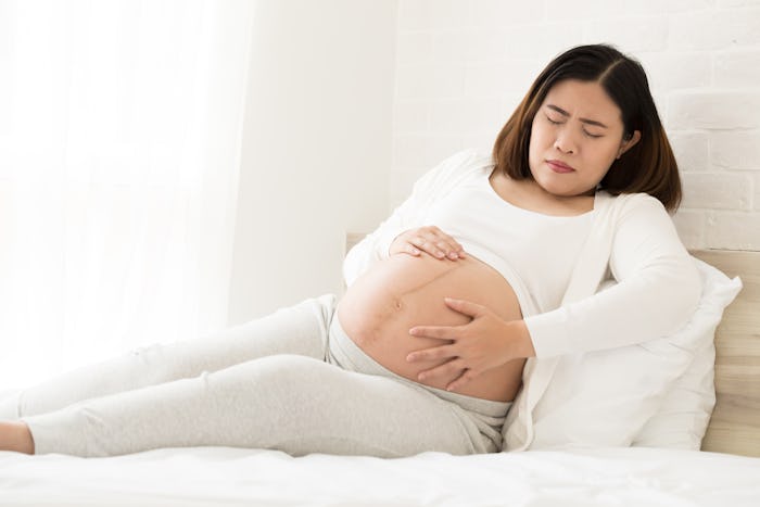 pregnant woman laying on white bed uncomfortably holding belly with both hands
