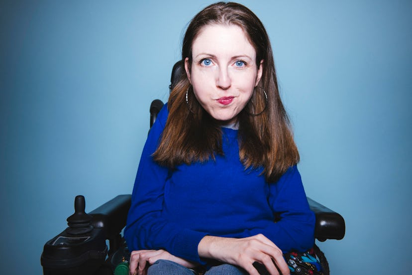 A woman in a blue shirt sits in a wheel chair with her arms crossed looking straight at the camera
