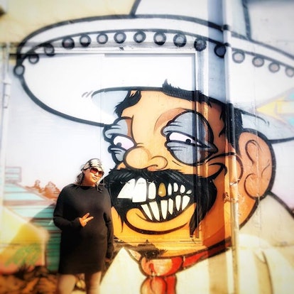 Celeste Winkel, an Asian-Latinx woman standing in front of graffiti with an illustrated Mexican man ...