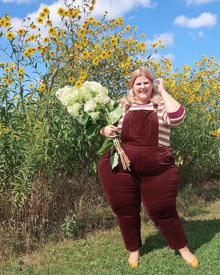 Plus-Size Influencer Anna O'Brien wearing burgundy cord overalls and holding flowers while posing ou...