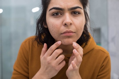 A woman wearing a brown hoodie looks in the mirror and checks the acne on her chin