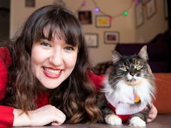 A happy brunette woman in red lipstick and a red sweater poses with her cat on the floor for a pictu...