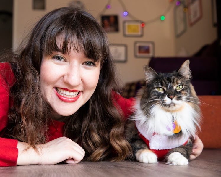 A happy brunette woman in red lipstick and a red sweater poses with her cat on the floor for a pictu...