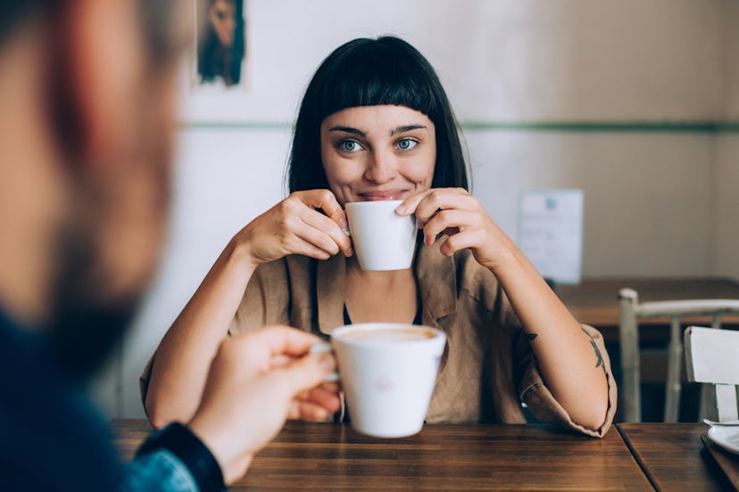 A happy woman smiles and holds up her cup of coffee while looking at her partner across the table.