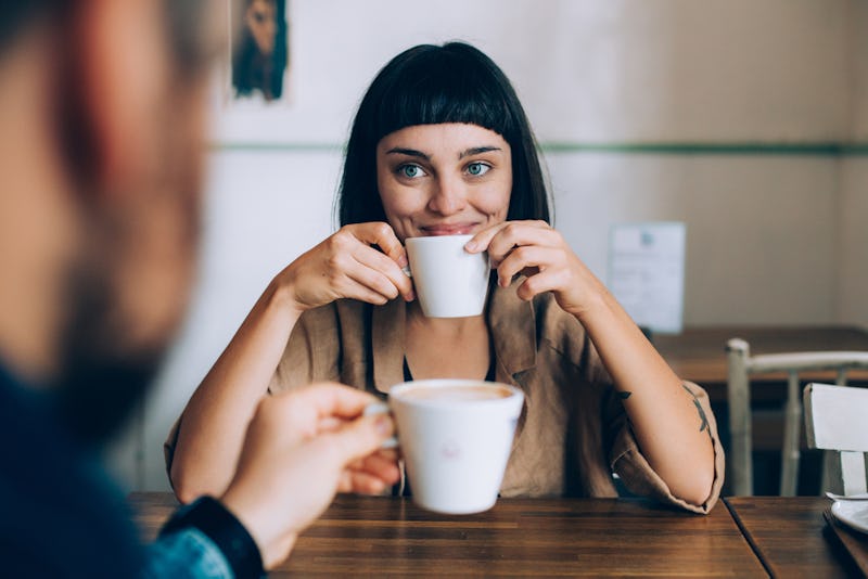 A happy woman smiles and holds up her cup of coffee while looking at her partner across the table.