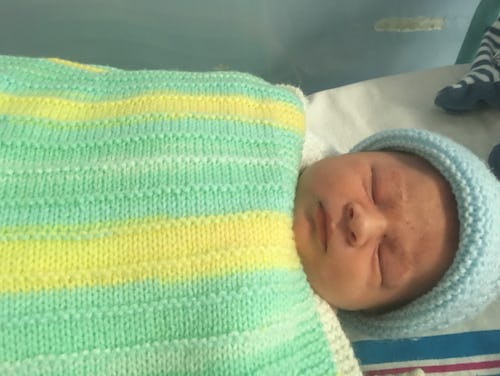 A newborn baby sleeping while wearing a knitted blue hat and covered by a knitted blanket