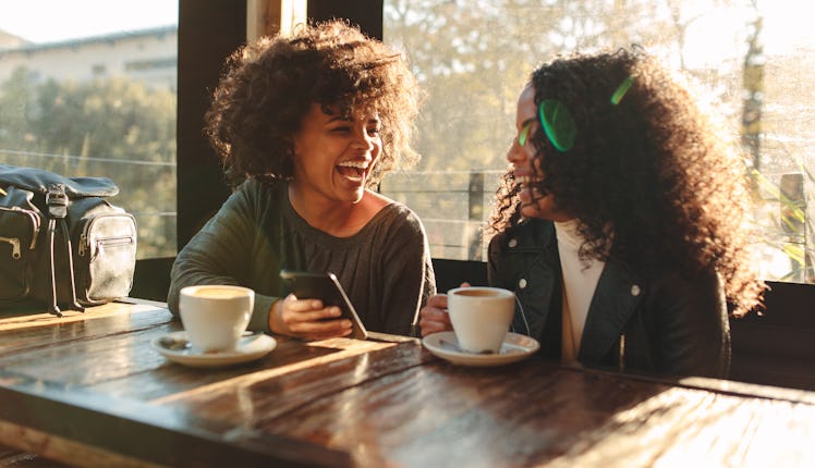 Two friends laugh at a coffee shop on a sunny day, while one of them holds a cellphone and the other...