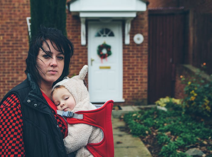 A mother holding her baby in a baby carrier in front of her red brick house