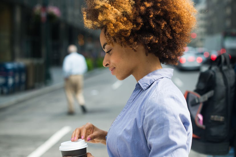A woman with an afro, in a blue button-up shirt walking down the street with a cup of coffee