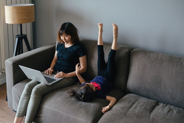 A mother working on her laptop while sitting on a couch and her daughter playing next to her