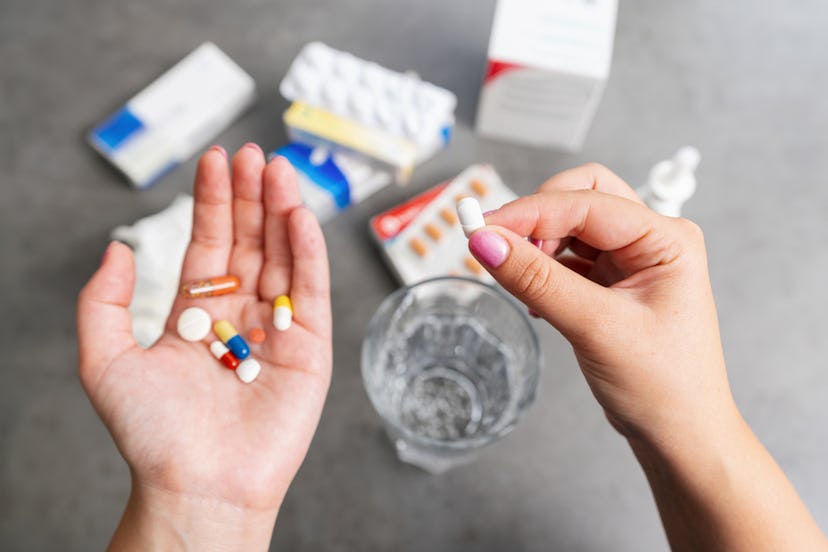 A woman taking 8 different pills with pill packages and a glass of water on a grey surface