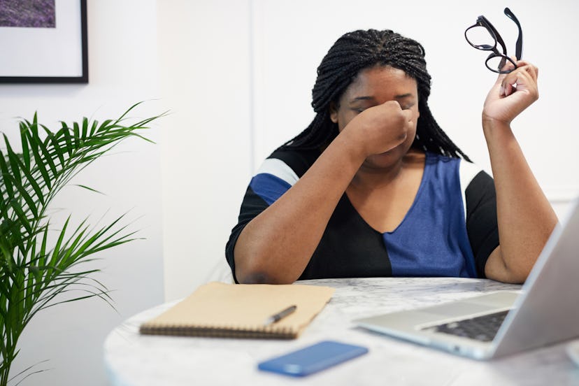 A woman who has taken of her glasses to rub her eyes white her laptop notebook and phone in front of...