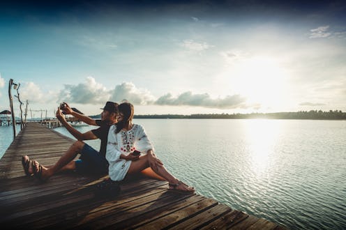A couple on a Valentine’s Day Weekend Getaway  taking a selfie while sitting on a pier with the sea ...
