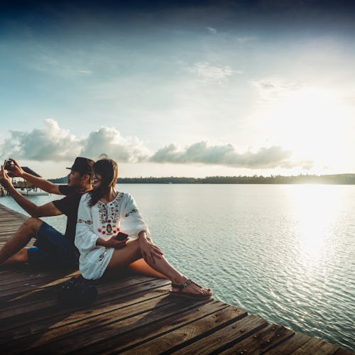 A couple on a Valentine’s Day Weekend Getaway  taking a selfie while sitting on a pier with the sea ...