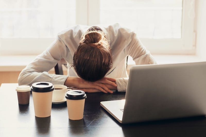 A woman in a difficult work environment sitting with her head down in front of a laptop and coffee c...