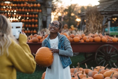A girl in a white dress and jean jacket is posing for her best friend in a pumpkin patch for Hallowe...