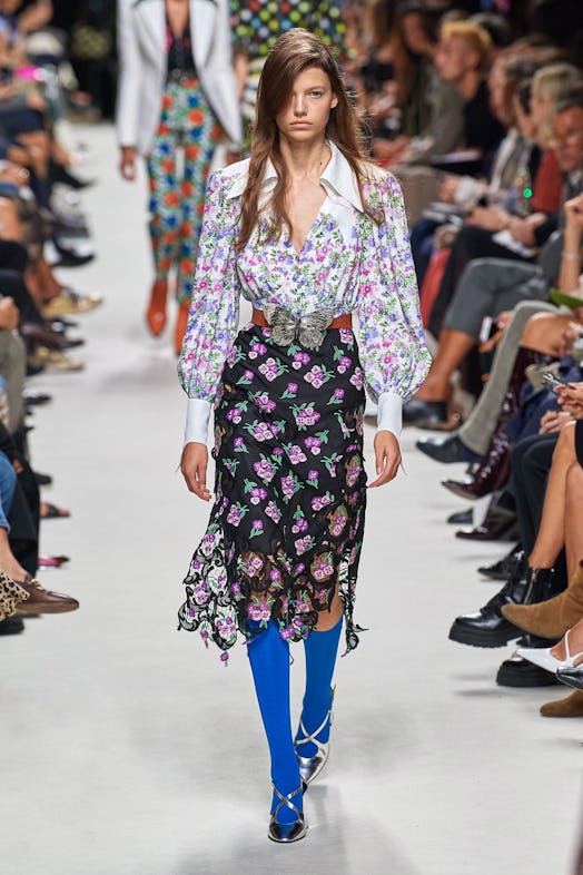 a model wearing a patterned button up shirt and a skirt with blue leggings down the runway