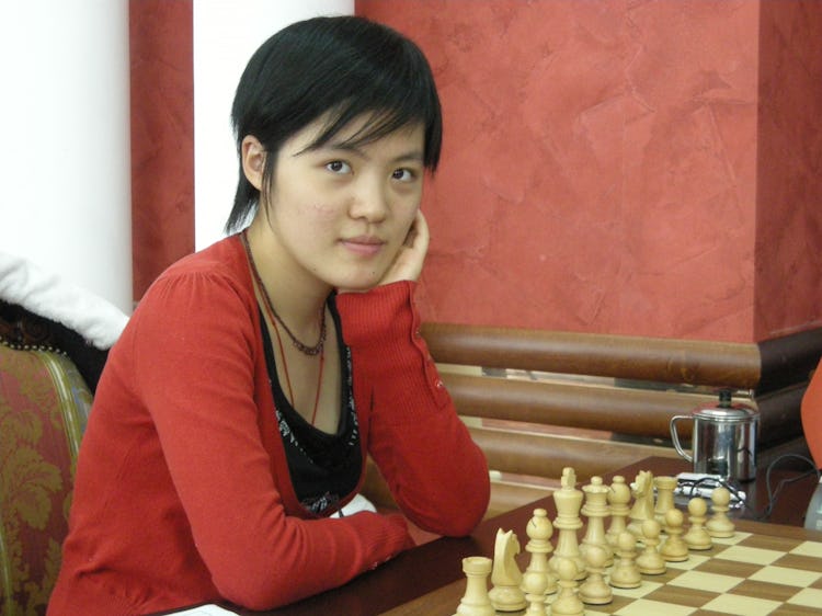 Girl in red sweater sits with arms crossed at chess table.