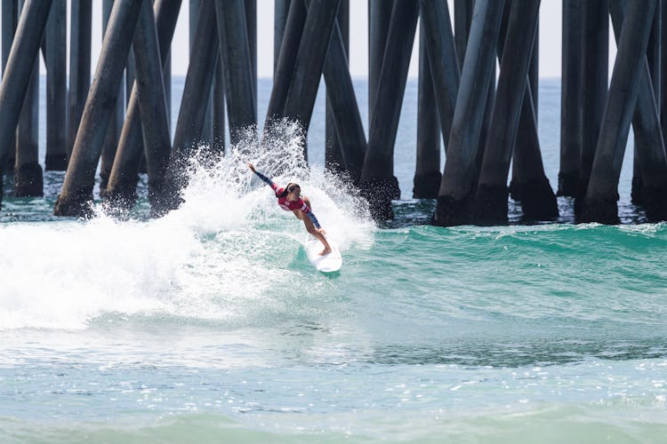 A teenaged surfer balances on top of a wave by a pier.