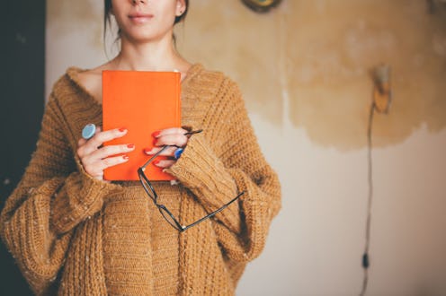 A woman in a sweater clutches an orange hardcover book, in this list of horror novels to read, based...