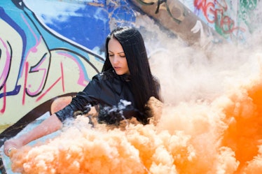A woman stands next to a wall covered in graffiti in the city with orange smoke around her.