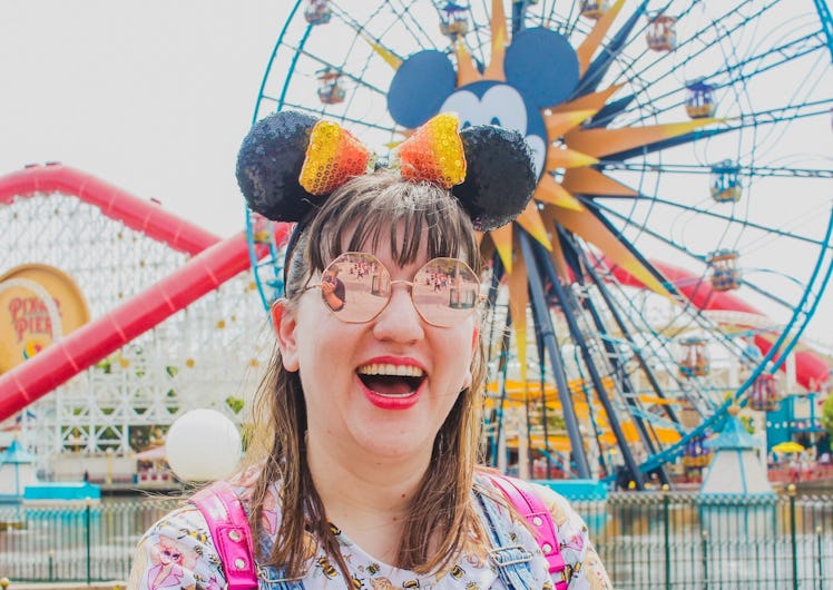 A woman laughs at the camera in a pair of sunglasses, Minnie ears, and a hot pink backpack in front ...