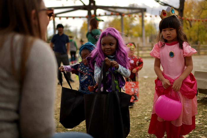 a girl in a purple wig and a girl dressed as a pink princess hold out pumpkin baskets for candy on H...