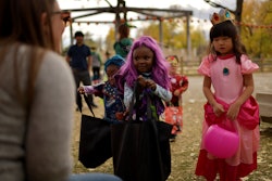 a girl in a purple wig and a girl dressed as a pink princess hold out pumpkin baskets for candy on H...