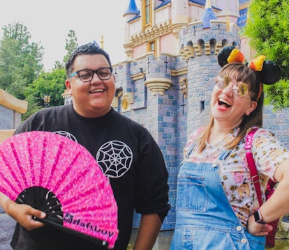 Two friends are standing in front of the Disneyland castle, one holding a pink fan and the other smi...