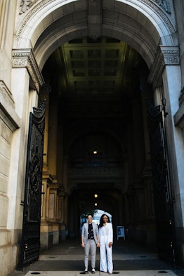 A newly-married couple is standing under an arch in the city in chic clothes and holding hands, smil...