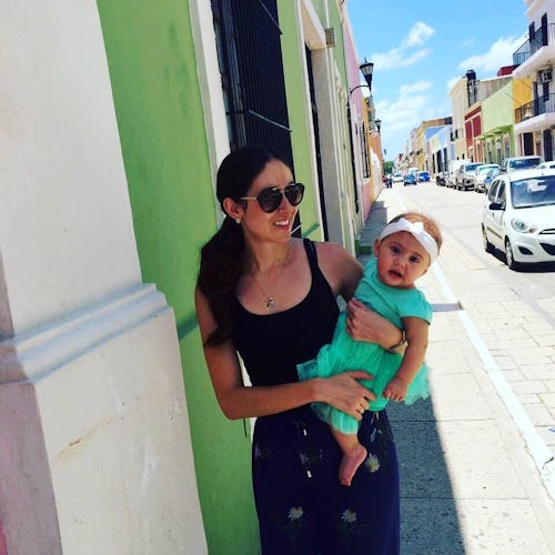 A woman stands on street in Mexico against green wall holding a baby.