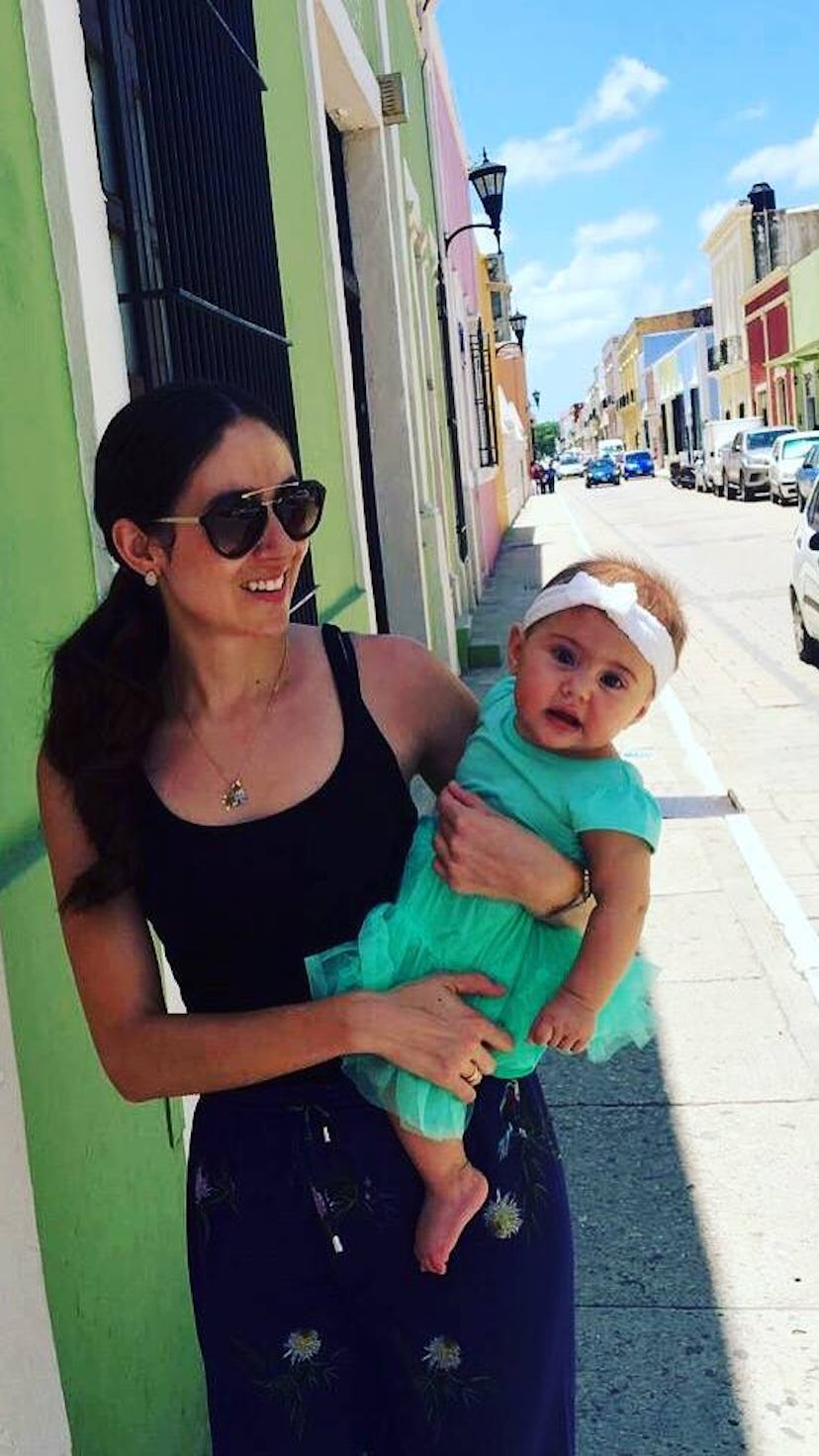 A woman stands on street in Mexico against green wall holding a baby.