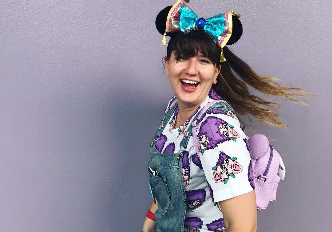 A woman smiles in front of a purple wall, wearing overalls, Minnie ears, and a purple backpack at Di...