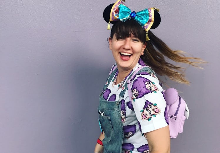 A woman smiles in front of a purple wall, wearing overalls, Minnie ears, and a purple backpack at Di...