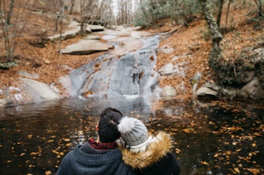 A couple cuddles and sits in front of a small waterfall in a forest with lots of fall foliage for le...
