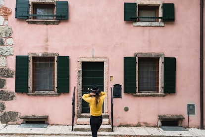A stylish girl in a yellow sweater and black felt hat stands in front of a pink, rustic building.