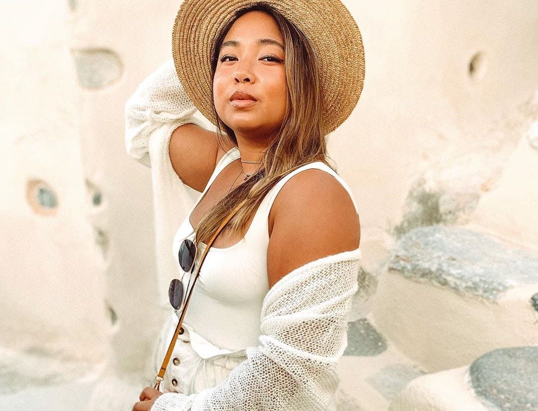 A girl who's posing in front of a while wall wearing a tan hat, white sweater, and white tank top ha...