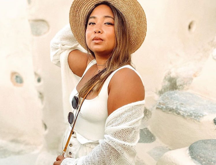 A girl who's posing in front of a while wall wearing a tan hat, white sweater, and white tank top ha...